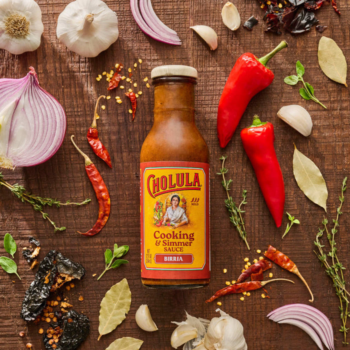 A bottle of Cholula Birria Cooking &amp; Simmer Sauce is surrounded by ingredients like garlic red onion red peppers and bay leaves on a wooden surface.