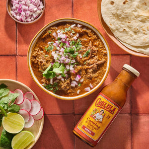 A bowl of Birria topped with red onions and cilantro accompanied by tortillas radishes limes and a bottle of Cholula Cooking &amp; Simmer Sauce.