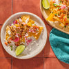 A plate of nachos topped with pulled pork pico de gallo and lime wedges sits next to another plate of nachos and a teal napkin.