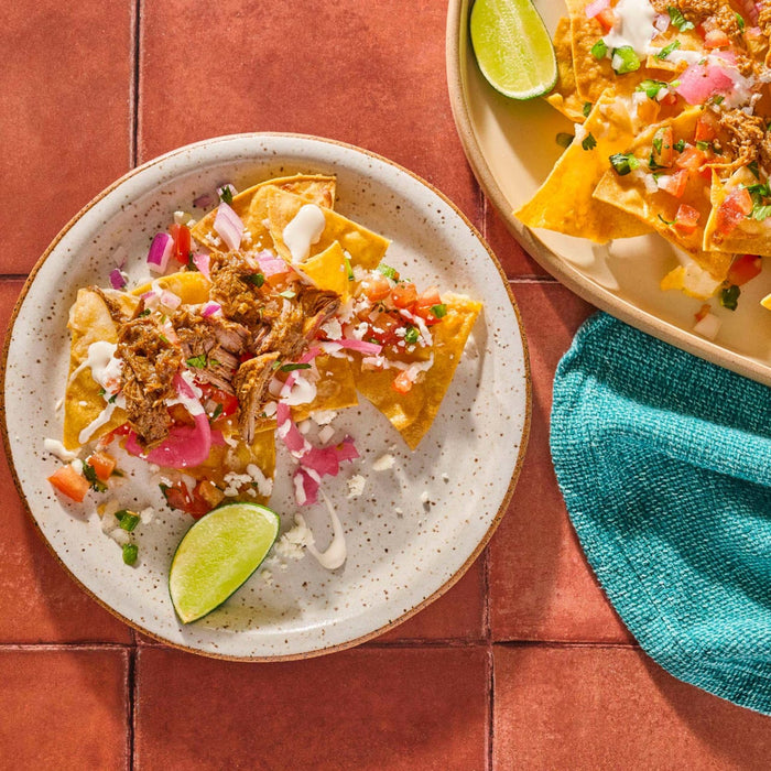 A plate of nachos topped with pulled pork pico de gallo and lime wedges sits next to another plate of nachos and a teal napkin.