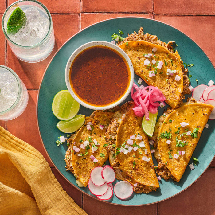 Overhead shot of a plate of birria tacos with dipping sauce lime wedges radishes and pickled onions accompanied by two glasses of a clear beverage with lime.