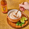 A hand stirs a bowl of creamy sauce with Cholula Topping &amp; Cooking Sauce accompanied by lime wedges and cilantro.