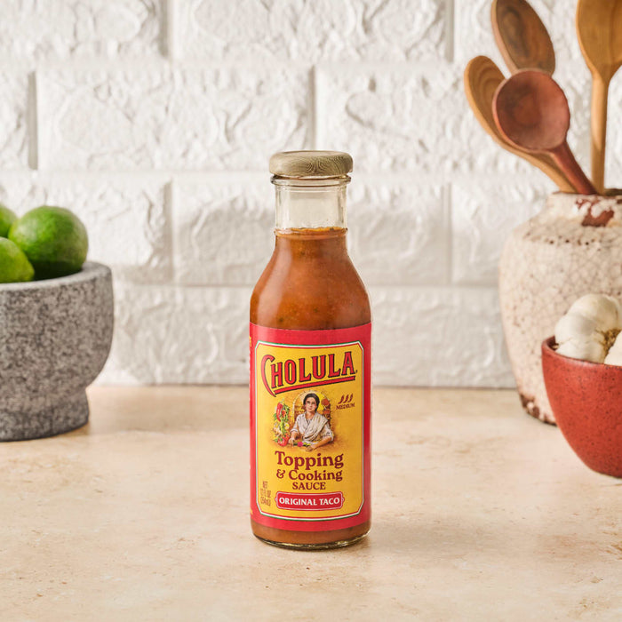 A bottle of Cholula Original Taco Topping &amp; Cooking Sauce sits on a counter with limes garlic and wooden spoons in the background.