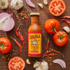A bottle of Cholula Original Taco Topping &amp; Cooking Sauce is surrounded by fresh ingredients like tomatoes onions garlic and dried chili peppers on a wooden surface.
