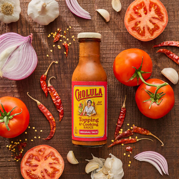A bottle of Cholula Original Taco Topping &amp; Cooking Sauce is surrounded by fresh ingredients like tomatoes onions garlic and dried chili peppers on a wooden surface.