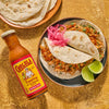 A bottle of Cholula Original Taco Topping &amp; Cooking Sauce is displayed next to a plate of tacos with pickled onions and lime wedges along with a stack of tortillas.