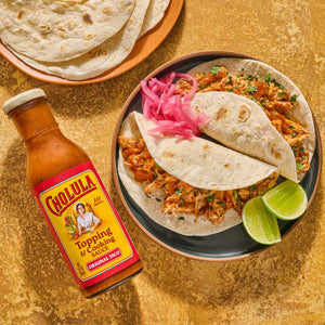 A bottle of Cholula Original Taco Topping &amp; Cooking Sauce is displayed next to a plate of tacos with pickled onions and lime wedges along with a stack of tortillas.