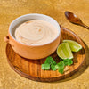 A bowl of creamy soup is garnished with lime wedges and cilantro on a wooden plate accompanied by a wooden spoon.