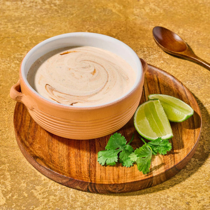 A bowl of creamy soup is garnished with lime wedges and cilantro on a wooden plate accompanied by a wooden spoon.