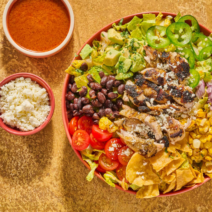 A vibrant and appetizing salad featuring grilled chicken black beans avocado corn tomatoes tortilla chips and jalapenos served with a side of dressing and crumbled cheese.