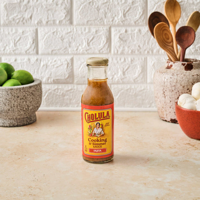 A bottle of Cholula Fajita Cooking &amp; Simmer Sauce sits on a countertop with limes wooden spoons and garlic in the background.