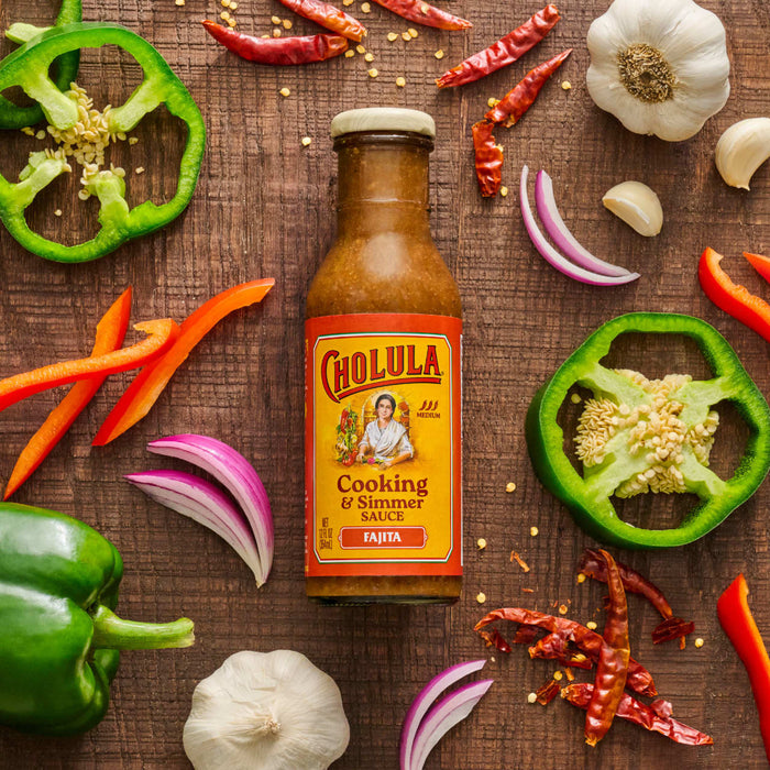 A bottle of Cholula Fajita Cooking &amp; Simmer Sauce is surrounded by fresh ingredients like peppers garlic and onions on a wooden surface.