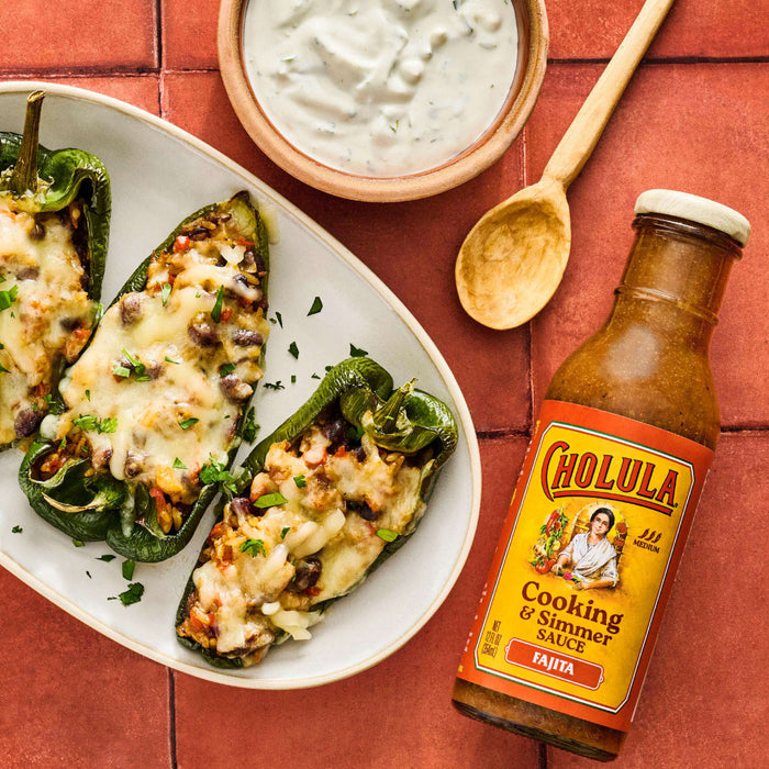 This is a food photography featuring stuffed poblano peppers on a plate a bowl of white sauce a wooden spoon and a bottle of Cholula Fajita Cooking &amp; Simmer Sauce.