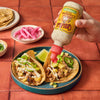 A hand pours Cholula Cremosa Cilantro Lime sauce onto two tacos filled with meat pineapple and jalapeA?os.
