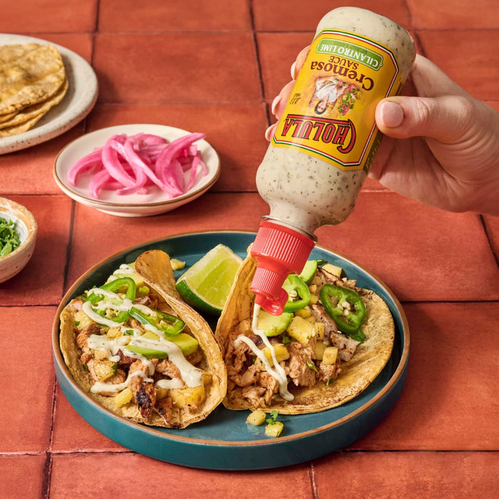 A hand pours Cholula Cremosa Cilantro Lime sauce onto two tacos filled with meat pineapple and jalapeA?os.