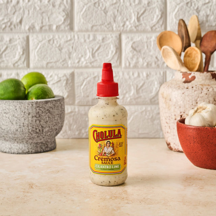 A bottle of Cholula Cremosa Cilantro Lime sauce sits on a counter with limes garlic and wooden spoons in the background.