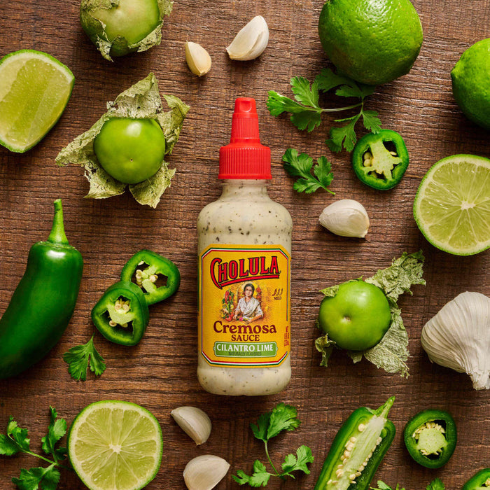 A bottle of Cholula Cremosa Cilantro Lime sauce is surrounded by fresh ingredients like limes tomatillos jalapeA?os garlic and cilantro.