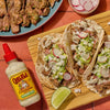Cholula Cremosa Sauce Cilantro Lime is featured alongside a wooden board with three tacos filled with steak guacamole radishes onions and sauce.