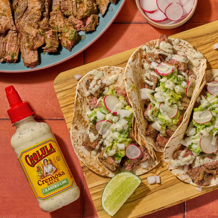 Cholula Cremosa Sauce Cilantro Lime is featured alongside a wooden board with three tacos filled with steak guacamole radishes onions and sauce.