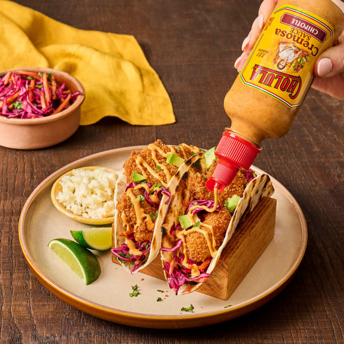 A hand pours Cholula Cremosa Chipotle sauce onto two tacos filled with fried food, red cabbage, and avocado, served with lime wedges and a side of cheese.