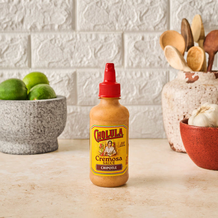 A bottle of Cholula Cremosa Chipotle sauce sits on a counter with limes garlic and wooden spoons in the background.
