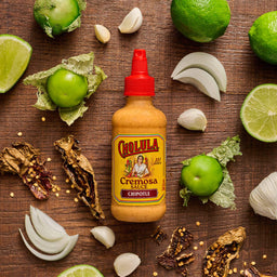 A bottle of Cholula Cremosa Chipotle sauce is surrounded by fresh ingredients like limes tomatillos garlic onions and dried chiles on a wooden surface.