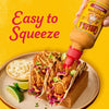 A bottle of Cholula Cremosa Chipotle sauce is being squeezed onto two tacos filled with fried food, red cabbage, and avocado, with a side of cheese and lime wedges.