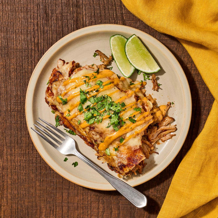Overhead shot of a plate of enchiladas topped with cheese sauce and cilantro accompanied by lime wedges and a fork set on a wooden table with a yellow napkin.