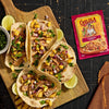 A wooden board displays four tacos topped with mango salsa and cilantro, alongside lime wedges and a packet of GOYA Sazon seasoning.