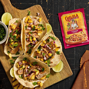 A wooden board displays four tacos topped with mango salsa and cilantro, alongside lime wedges and a packet of GOYA Sazon seasoning.