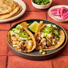 Two tacos al pastor with pineapple, jalapeño, and a lime wedge are displayed on a plate, accompanied by tortillas, cilantro, and pickled onions.