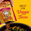 A hand pours Cholula taco seasoning into a pan of vegetables, highlighting its use for veggie tacos.