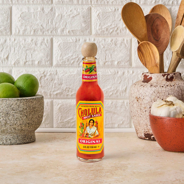 A bottle of Cholula Original Hot Sauce 5 Fl Oz stands prominently on a kitchen counter, flanked by fresh limes in a stone bowl and wooden cooking utensils in a decorative ceramic vase, with garlic cloves in a small red bowl nearby, all against a white brick-patterned wall.