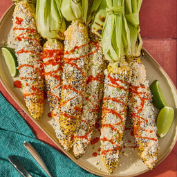 Grilled elote, or Mexican street corn, on a platter, generously coated with white cheese, green herbs, and drizzled with Cholula Original Hot Sauce 5 Fl Oz, served with lime wedges.