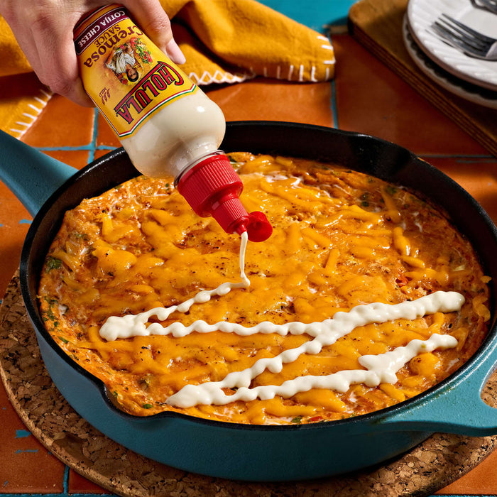 A person drizzles a bottle of Yellowbird Habanero Condiment over a cheesy casserole in a skillet.
