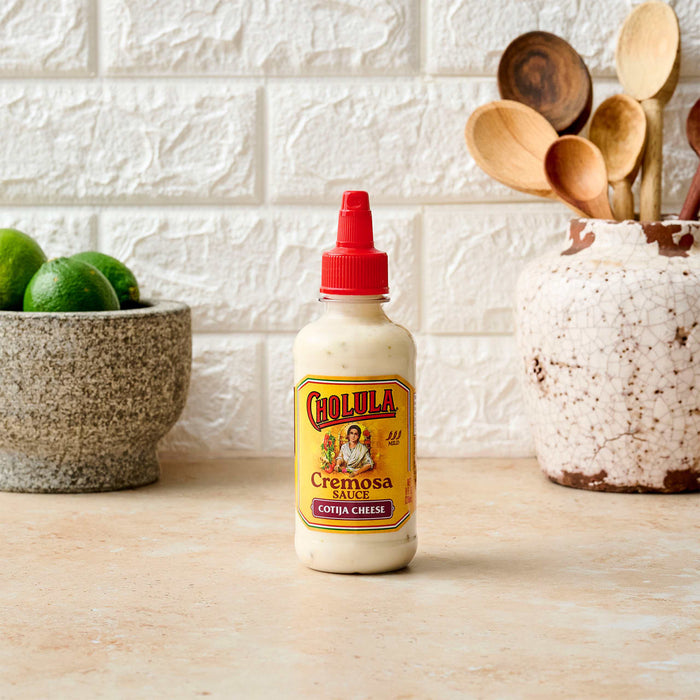 A bottle of Cholula Creamy Chipotle sauce sits on a countertop next to limes and kitchen utensils.