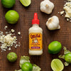 A bottle of Cholula Creamy Poblano sauce is surrounded by limes, garlic, and grated cheese on a wooden surface.