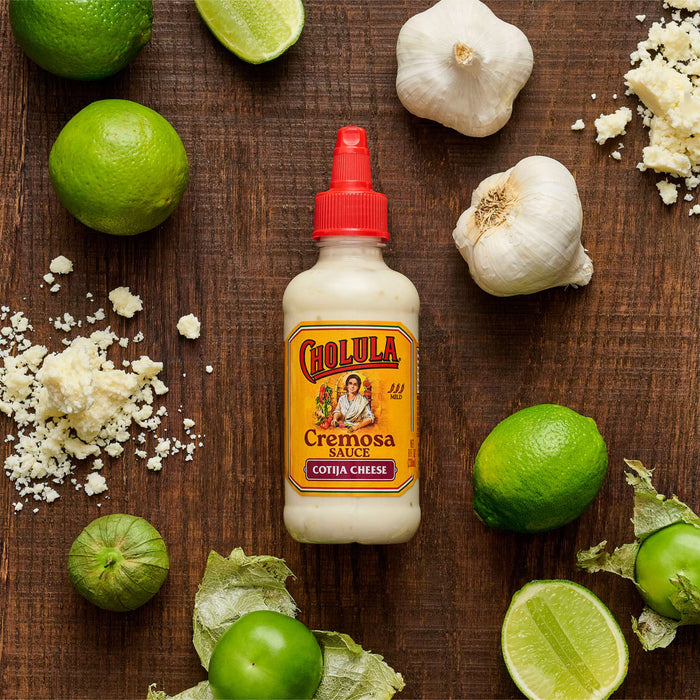 A bottle of Cholula Creamy Poblano sauce is surrounded by limes, garlic, and grated cheese on a wooden surface.