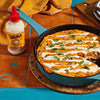 A skillet of Cholula Creamy Jalapeño Taco Dip is displayed on a colorful tile surface, with a bottle of Cholula Creamy Jalapeño sauce beside it.