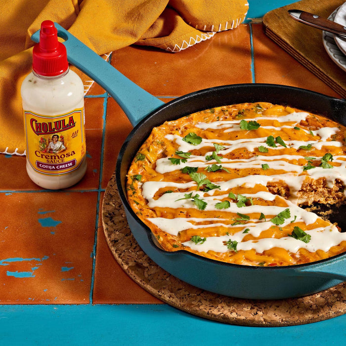 A skillet of Cholula Creamy Jalapeño Taco Dip is displayed on a colorful tile surface, with a bottle of Cholula Creamy Jalapeño sauce beside it.