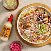 A vibrant bowl featuring grilled chicken, black beans, mango, pickled onions, and cotija cheese, drizzled with Cholula Creamy Jalapeño sauce, alongside a bottle of the sauce.