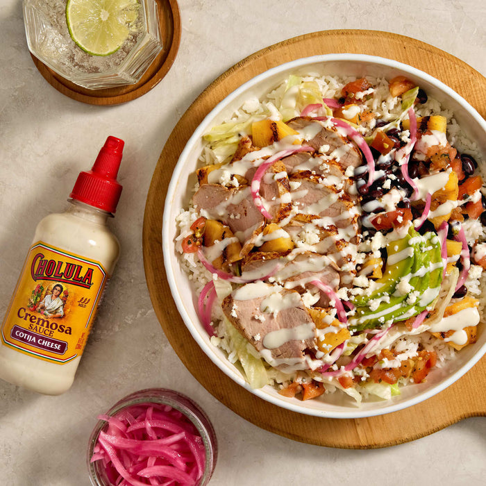A vibrant bowl featuring grilled chicken, black beans, mango, pickled onions, and cotija cheese, drizzled with Cholula Creamy Jalapeño sauce, alongside a bottle of the sauce.