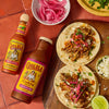 A bottle of Cholula Original Hot Sauce 5 Fl Oz and a bottle of Cholula Cooking Sauce Cochinita Pibil are displayed on a reddish-brown tiled surface next to two tacos topped with shredded meat, pickled red onions, and crumbled cheese.