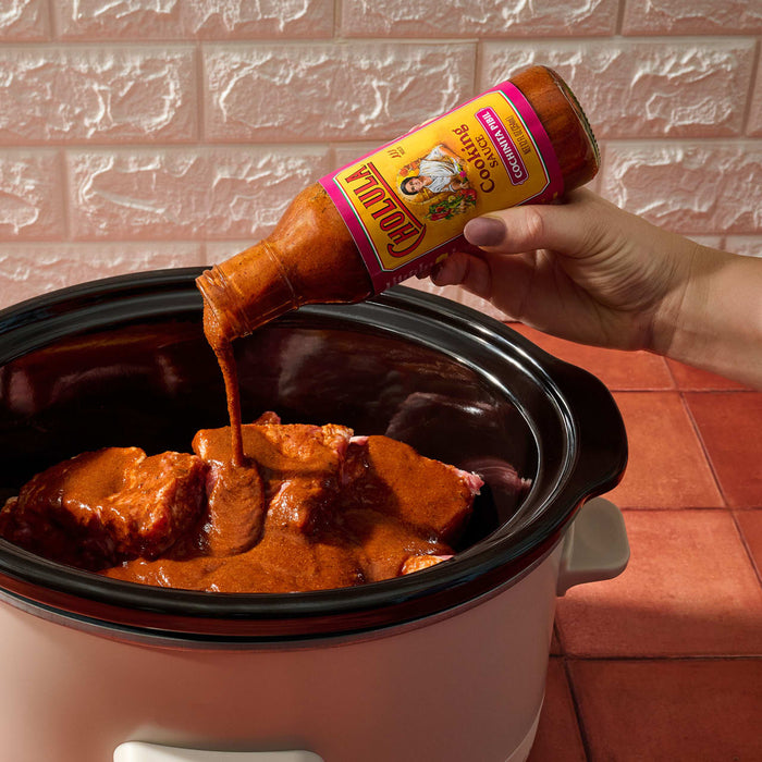 A hand pours Cholula Cocinita Pibil Cooking Sauce over meat in a slow cooker.