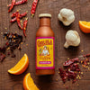 A bottle of Cholula Cochinita Pibil Cooking Sauce is surrounded by garlic cloves, orange slices, and dried red peppers on a wooden surface.
