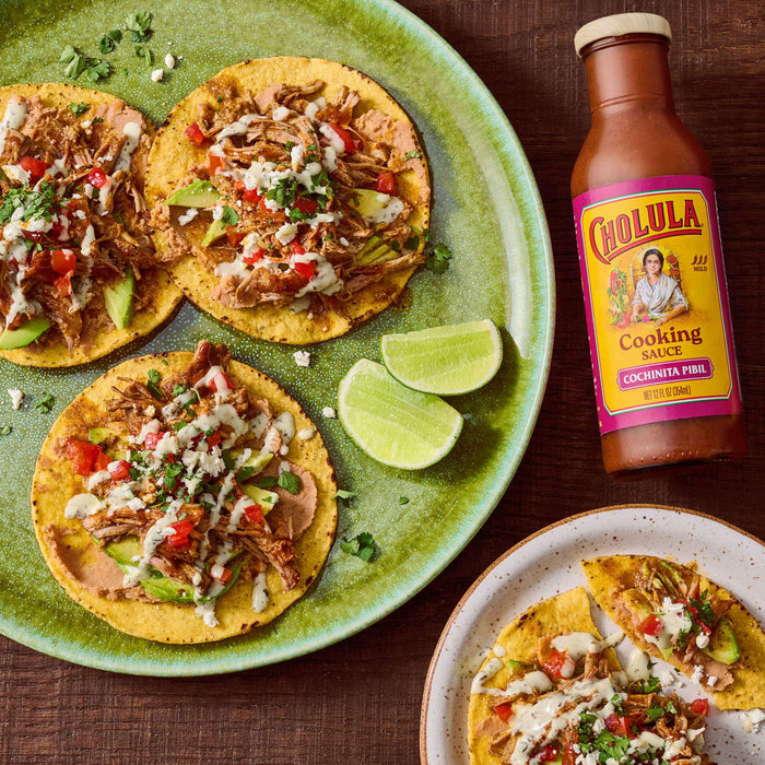 Overhead shot of tostadas topped with pulled pork, avocado, tomatoes, and cheese, accompanied by lime wedges and a bottle of Cholula Cooking Sauce Cochinita Pibil.