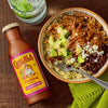 A bottle of Cholula Cooking Sauce Cochinita Pibil is next to a bowl of rice, beans, avocado, and shredded meat, with a lime drink in the background.