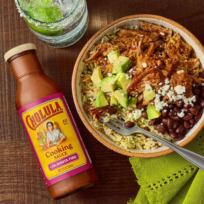 A bottle of Cholula Cooking Sauce Cochinita Pibil is next to a bowl of rice, beans, avocado, and shredded meat, with a lime drink in the background.