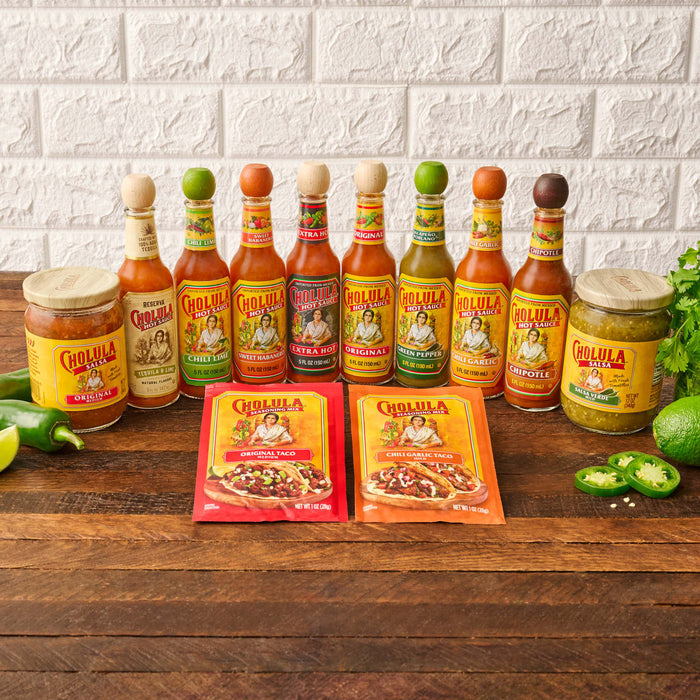 A variety of Cholula brand products, including several bottles of Cholula Original Hot Sauce, other hot sauce flavors, salsas, and seasoning mixes, are displayed on a rustic wooden table with fresh ingredients.