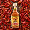 A bottle of Cholula Original Hot Sauce, featuring its iconic wooden cap, is centered on a dark wooden surface, surrounded by a vibrant array of dried red chili peppers and scattered seeds.